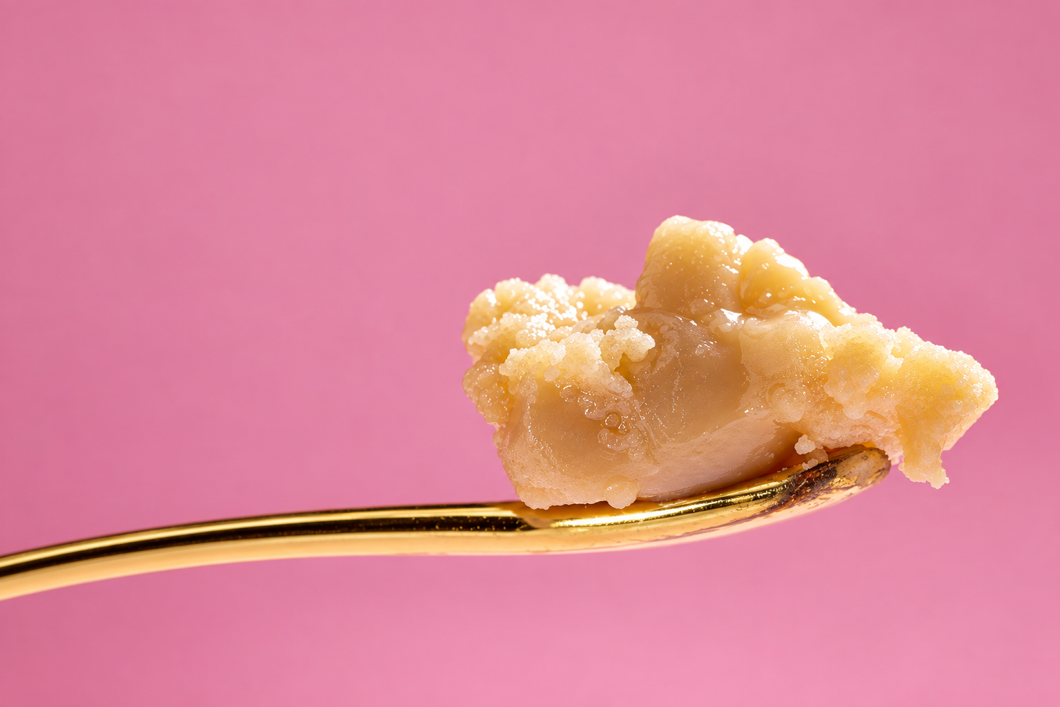 Close-up of warm golden honey with fine, softly folded texture on a polished gold tool against a seamless pink background